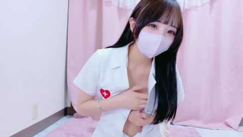 Media: Video of an East Asian woman in a white nurse outfit, wearing a white mask, sitting on a pink bed with soft pink curtains. Her long black hair hangs down, and she has a gentle, shy expression.