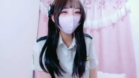 Media: Video of a young Asian woman with long black hair, wearing a white mask, a light blue shirt, and a green hat, in a soft pink room with a floral curtain.