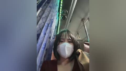Video of an Asian woman with straight black hair, wearing a white surgical mask, sitting on an airplane, with blue and green patterned curtains, and a beige jacket.