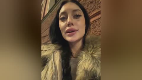 Media: Video of a young woman with long dark hair, wearing a fur coat, standing against a brick wall, slightly out of focus.