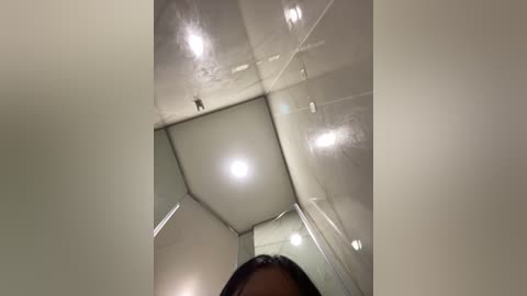 Media: Video of a dark-skinned person, likely male, standing in a small, narrow, white-tiled shower room with a ceiling light reflecting on the glossy tiles.