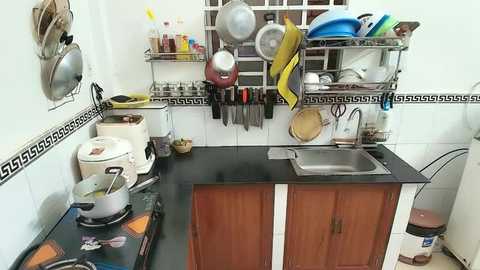 A video of a small, cluttered kitchen with a black countertop, wooden cabinets, stainless steel sink, and hanging utensils on a white tiled wall. The scene is chaotic with various kitchen items, including a rice cooker, pots, and utensils.