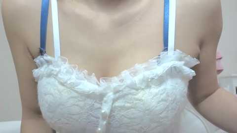 Media: Video of a fair-skinned woman wearing a white lace bra with blue straps, ruffled edges, and a bow in the center, highlighting her medium-sized breasts.