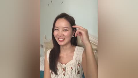 Video of a smiling East Asian woman with long dark hair, wearing a white crochet top, in a softly lit bedroom with pale walls.