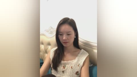 Video of a young Asian woman with long, straight black hair, wearing a beige crochet top, sitting on a bed with a cream tufted headboard. The background is plain and light-colored.