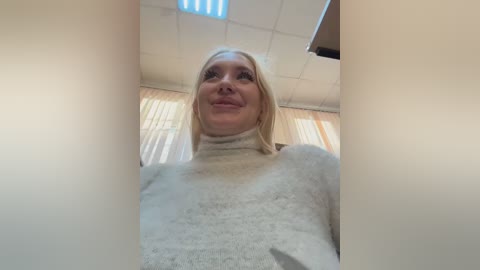 Media: A video of a smiling, blonde woman in a light-colored sweater, taken from a low angle, showing off her chest and shoulders. The background features a white ceiling and light-colored curtains.