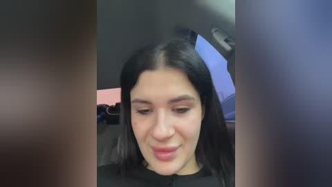 Video of a young woman with long black hair and fair skin, wearing a black top, seated in a dimly lit room with blurred background.