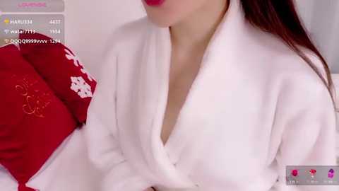 Video of a woman in a white, fluffy robe with a deep V-neck, lying on a bed with red pillows and a white duvet.