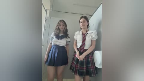 Video of two women in school uniforms, one in a navy-blue skirt, the other in a red plaid skirt, standing in a dimly lit restroom.