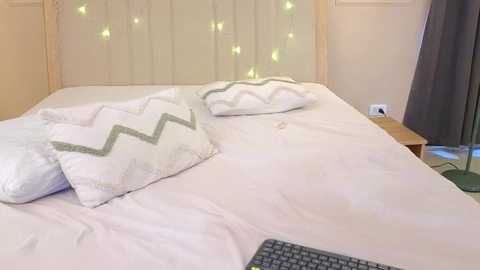Video of a neatly made bed with white linens, two white pillows with green zigzag patterns, and a black keyboard placed at the bottom. The background features beige walls, a wooden headboard, and fairy lights.