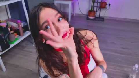 Video of a young woman with long dark hair, wearing a red dress, playfully covering her face, in a modern living room with wooden floors, shelves, and potted plants.