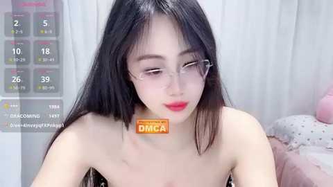 Media: A video of an Asian woman with long black hair, fair skin, and glasses, wearing a strapless black dress, in a bedroom with a pink teddy bear and a white bedsheet.