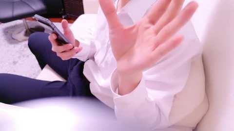 Video of a person in white business attire, with one hand raised in a stop gesture, using a smartphone while seated on a chair with a black leather cushion.