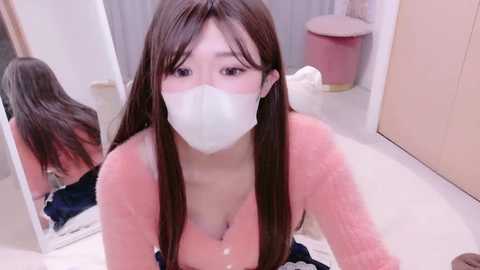 Video of an Asian woman with long brown hair, wearing a white mask and a pink shirt, kneeling in a bathroom, looking into a mirror, with a towel on the floor.