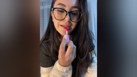 Video of a young woman with long dark hair and black-rimmed glasses, applying lipstick in a bathroom with white tiles and a window.