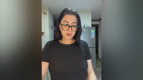 Video of a young woman with medium skin tone, long black hair, and black-framed glasses, wearing a tight black t-shirt, standing in a dimly lit hallway.