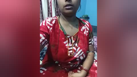 Video of a South Asian woman in a red saree with white geometric patterns, seated indoors. She wears a gold necklace, and the background includes patterned curtains and a blue wall.