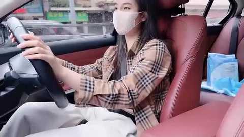 Media: Video of an Asian woman with long black hair, wearing a face mask, plaid shirt, and white pants, driving a red car with a blue bag on the seat.