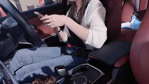 Media: Video of a woman with long brown hair, wearing a white shirt and blue jeans, seated in a car, reading a book while adjusting the seat controls. The car interior features red leather seats and a cluttered center console.