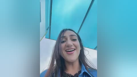 Video of a smiling Latina woman with long brown hair, wearing a blue denim shirt, standing in front of a glass window with turquoise tinted glass.