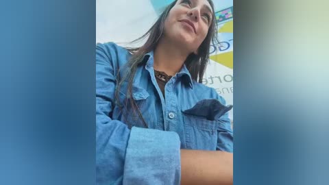 Video of a young woman with long dark hair, wearing a denim shirt, leaning back with a content expression, in an outdoor setting with a blue and yellow backdrop.