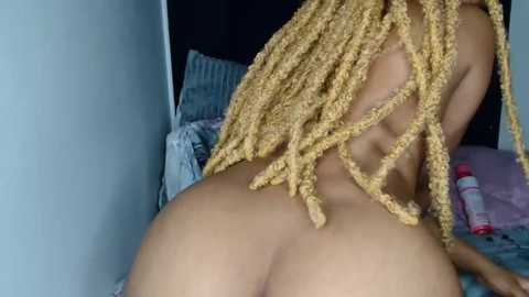 Video of a nude woman with light brown skin, long, blonde dreadlocks, and a curvy physique, kneeling on a bed.