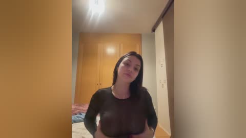 Video of a young woman with long black hair, wearing a black long-sleeved top, standing in a narrow hallway with beige walls, a wooden door, and a bed with a pink blanket visible in the background.