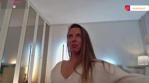 Video of a blonde woman with long hair, wearing a white blouse, standing in a dimly lit bedroom with mirrored wardrobes and a bedside lamp.