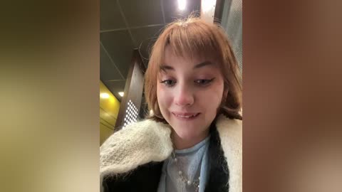 Media: Video of a young Caucasian woman with light brown hair and bangs, smiling, wearing a light blue shirt and a black and white knit sweater. She is in a fluorescent-lit room with tiled walls and a window.
