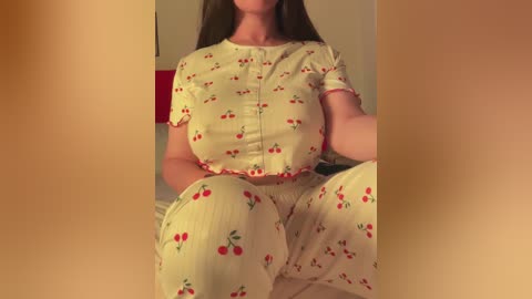 Video of a woman with long brown hair, wearing a yellow cherry-print nightgown and matching pajama pants, sitting on a bed. Warm lighting.
