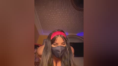 Media: Video of a woman with long, dark hair, wearing a red headband, black face mask, and a dark top, kneeling in a dimly-lit room with a textured ceiling and exposed fan.