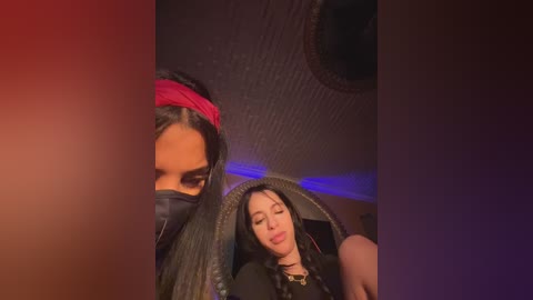 Video of a young woman with long black hair and a red headband, wearing a black face mask, seated in a dimly lit room with a tiled ceiling.