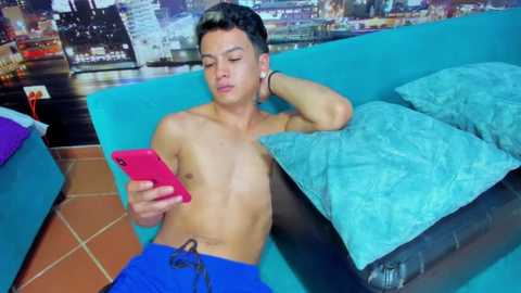 Media: Video of a shirtless young man with short dark hair, wearing blue shorts, lying on a teal sofa with a blue cushion, texting on a pink phone, tiled floor, and cityscape wallpaper background.