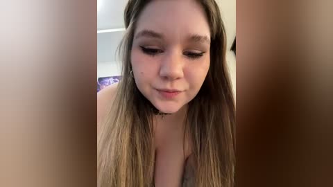 Media: Video of a young, fair-skinned woman with long, straight brown hair, wearing a white tank top, smiling slightly. The background is blurred, showing a bedroom with a bed.