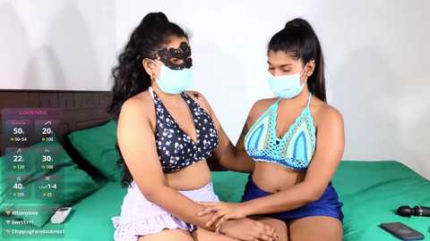 Video of two young South Asian women in matching blue face masks, sitting on a green bed. One wears a black lace mask, a black halter top, and white shorts; the other a blue halter top and blue shorts.