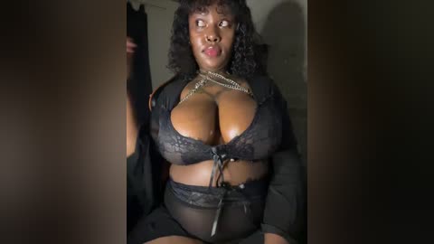 A video of a plus-sized Black woman with large breasts, wearing black lace lingerie and a necklace, standing in a dimly lit room.