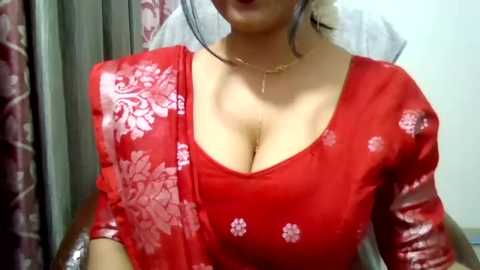 Media: Video of a light-skinned woman with a medium build, wearing a red sari with white floral patterns. She has a gold necklace and is seated indoors with maroon curtains in the background.