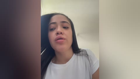 Video of a young woman with long black hair, light brown skin, and full lips, wearing a white t-shirt, captured in a bathroom with beige walls.