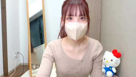 Video of an Asian woman with fair skin, red hair, and bangs, wearing a beige mask, beige long-sleeve top, and a Hello Kitty plush on the right. She sits in a room with a wooden door and a mirror in the background.