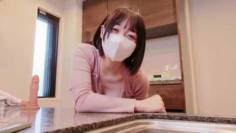 A video of an Asian woman with a pale complexion, short black hair, wearing a pink cardigan, white mask, and a large dildo on the kitchen counter.
