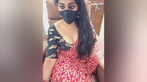 A video of a South Asian woman with medium brown skin, wearing a red and white floral sari with black mask, black short sleeves, and black mask, sitting in a tan chair indoors.