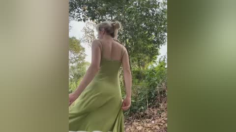Video of a fair-skinned woman with blonde hair in a messy bun, wearing a green, strapless, flowing dress, walking away from the camera in a lush, green forest setting.