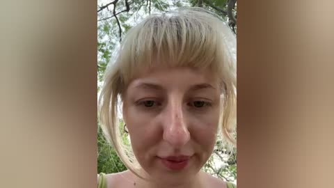 A video of a young Caucasian woman with short, blonde hair and bangs, wearing a green top, standing in a forest.