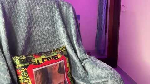 Media: Video of a woman's face on a green and yellow \"Che\" Guevara t-shirt, draped over a chair with a teal patterned cushion. Background features a partially visible door and wall with purple lighting.