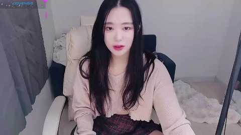 Video of an Asian woman with long black hair, fair skin, and red lipstick, wearing a beige sweater and plaid skirt, sitting in a cream leather chair with a fluffy white rug.