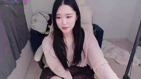 Video of an Asian woman with long black hair, fair skin, and light makeup, wearing a beige sweater and plaid skirt, sitting in a cluttered room with a mirror, a bed, and a fluffy rug.