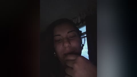 A dimly-lit video captures a woman with medium skin tone, wearing a black top, standing in a shadowy doorway. She has her hand near her mouth, suggesting contemplation or secrecy. The background shows a window with white curtains.