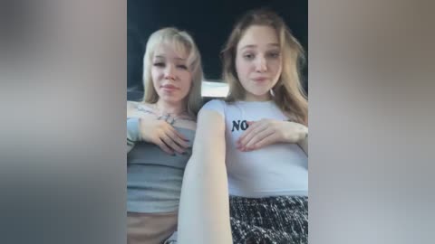 Media: Video of two young women, both with blonde hair, sitting in a car. One wears a grey crop top, the other a white shirt with \"NO\" on the chest.
