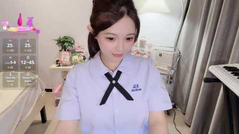 Media: Video of an East Asian woman in a white blouse with a black cross, sitting at a desk in a bright room with a whiteboard, calendar, and potted plants.