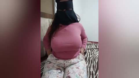 Media: Video of a woman with medium skin tone, wearing a black hijab, pink sweater, and zebra-print pants, sitting on a bed with a beige headboard in a dimly lit room.